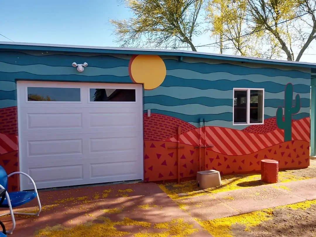 Colorful exterior mural with a yellow sun, blue sky bands, and layered red and orange desert forms beside a white garage door.