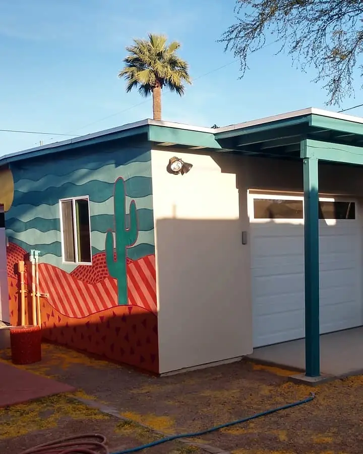 Garage and patio view showing a colorful desert landscape mural wrapping the building corner beneath a palm tree.