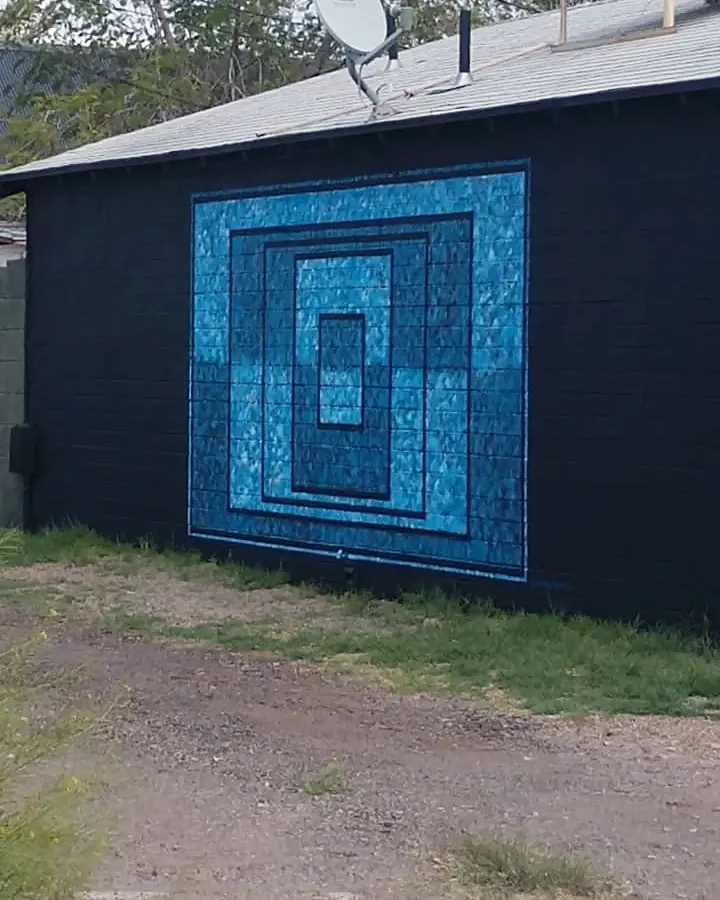 Blue geometric mural centered on a long black exterior wall beside a gravel drive and trees.