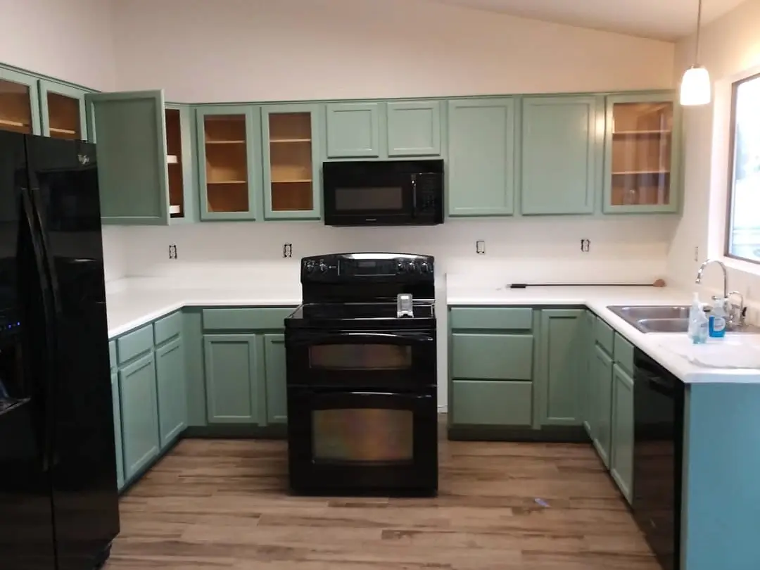 Kitchen after cabinet refinishing, with muted green cabinetry, white counters, and black appliances.