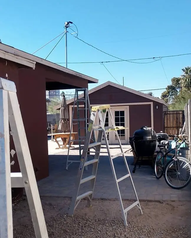Pale side wall and backyard structures before the navy exterior paint work, with a ladder on the jobsite.