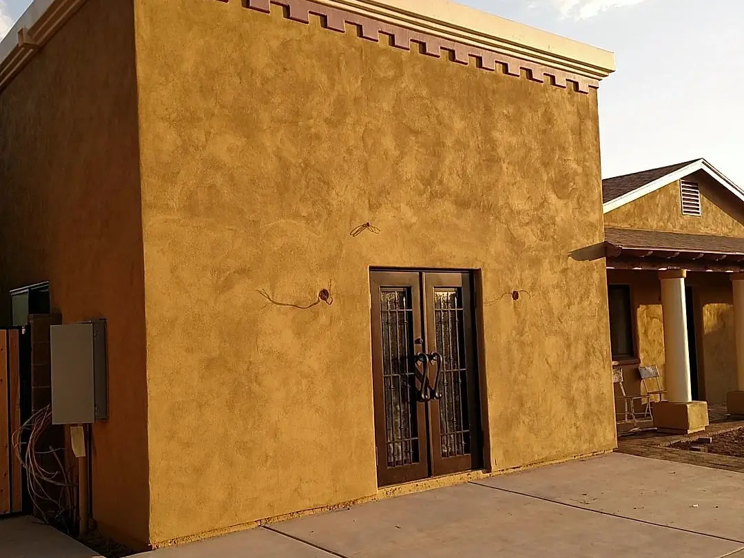 Mustard-colored stucco wall with a dark metal door and red geometric trim near the roofline.