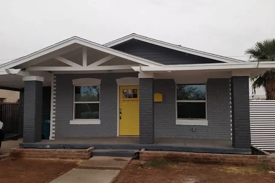Gray bungalow with white architectural trim and a bright yellow front door beneath a deep porch.