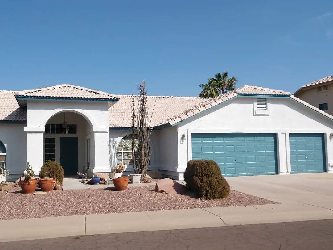 White stucco home with muted blue garage doors, a pale tiled roof, and desert landscaping under a clear sky.