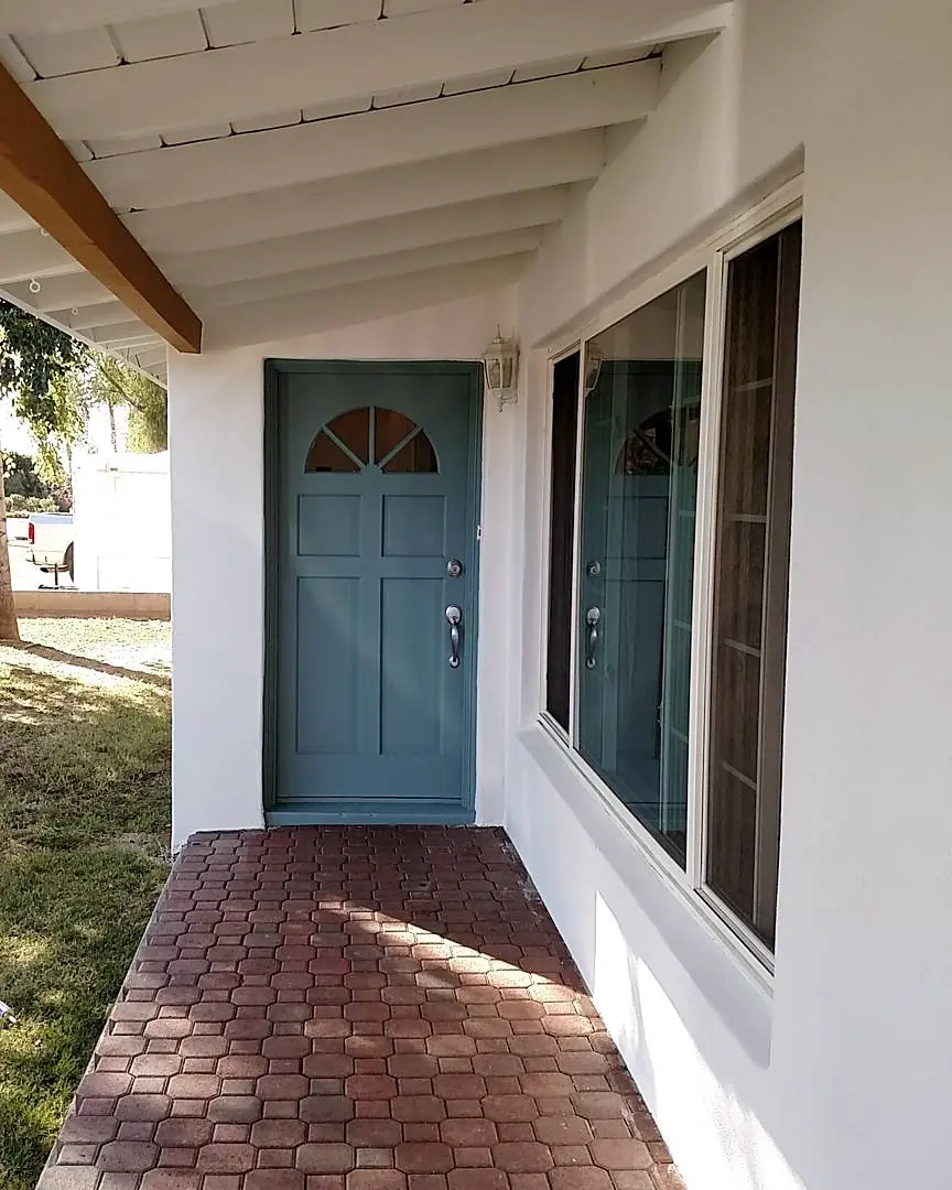 Blue front door set against bright white exterior walls beneath a shaded porch with a brick walkway.