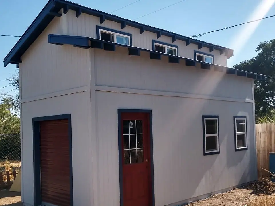 Finished backyard studio with light gray vertical siding, dark blue-gray trim, and a red door.
