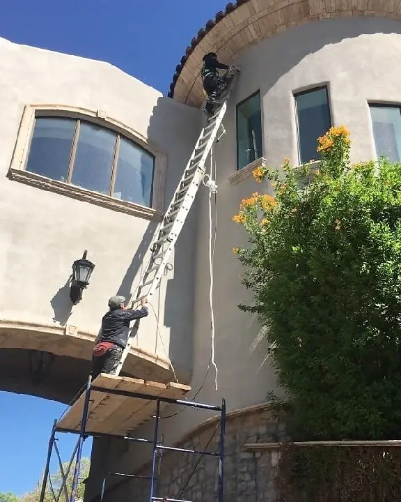 Two painters working from a ladder and scaffold on a curved stucco exterior below a tiled roof.